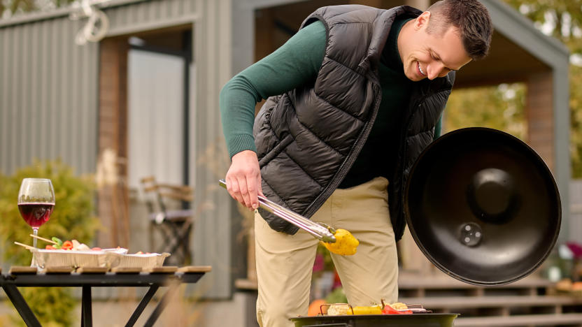 Smiling man leaning over the open brazier and gripping the slice of bell pepper with barbecue tongs