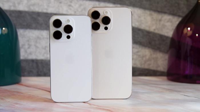 A white iPhone 16 Pro and a desert iPhone 16 Pro Max standing on a table.