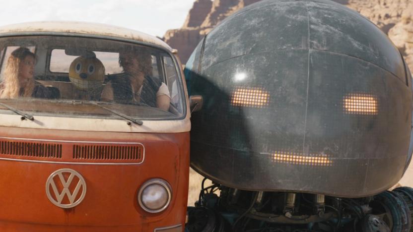 Two people sit inside a VW Bus alongside a robot with a yeallow head and an oversized smile. Next to the vehicle is the head of a much larger robot.